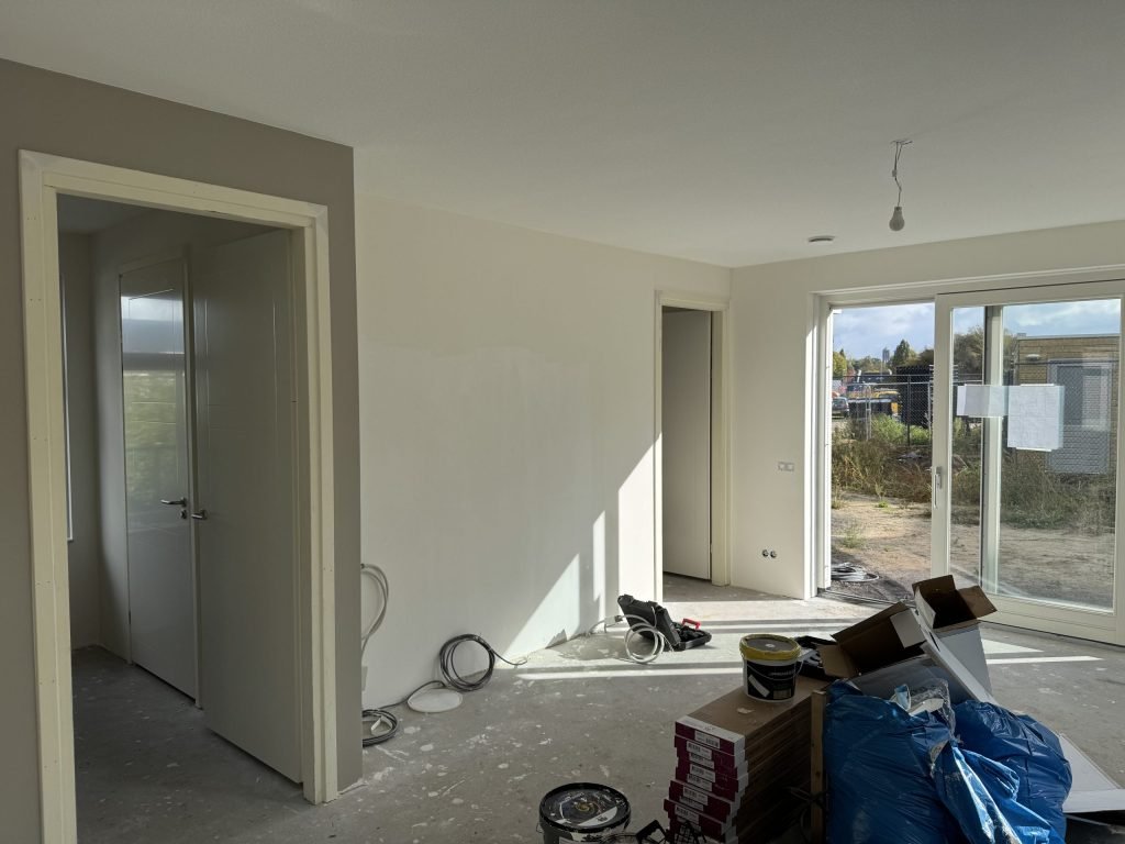 Open woonruimte van een net opgeleverde woning met lichte wanden, openstaande binnendeuren en een tuindeur naar het bouwterrein