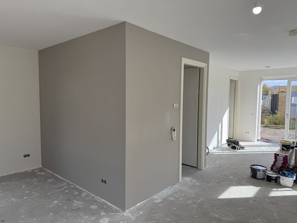 Grijs afgewerkte wand rond een kast in een nieuwbouwwoning, met verfemmers en pakken laminaat nog op de betonvloer