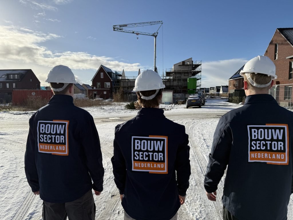 Drie medewerkers van Bouwsector Nederland in werkjas met logo kijken naar een nieuwbouwstraat met huizen in aanbouw en een kraan