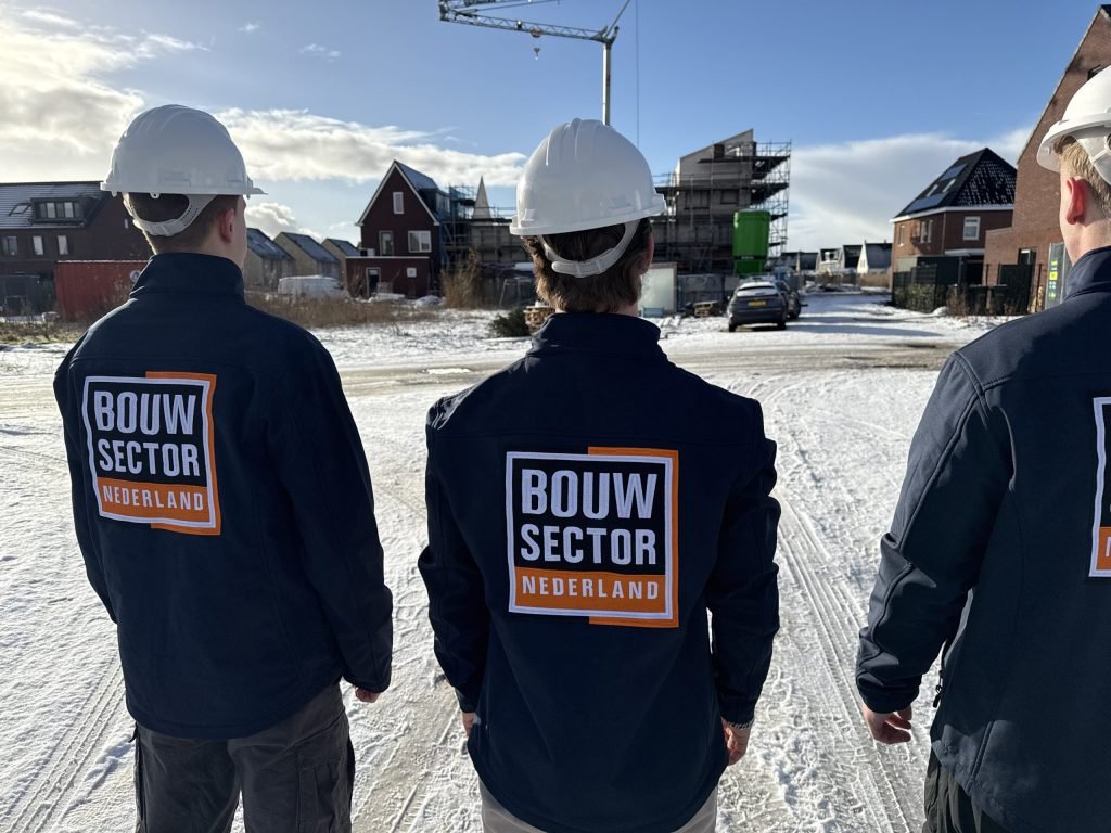 Drie vakmensen van Bouwsector Nederland met witte bouwhelm tussen nieuwe huizen in aanbouw en een bouwkraan in de sneeuw